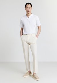Polo shirt bianco a maniche corte con texture a costine, pantaloni beige su misura, e scarpe slip-on beige, in piedi su uno sfondo neutro.
