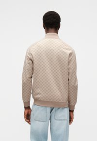 Blouson aviateur beige à motifs avec un tissu texturé, des poignets et un ourlet côtelés. Porté avec un jean bleu clair à coupe décontractée.