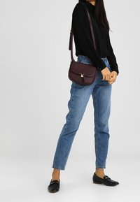 Žena v černém dlouhém rukávu, modrých džínách, černých mokasínech a nese malou tmavě fialovou crossbody tašku na jednobarevném pozadí.