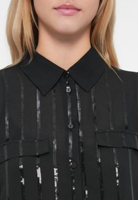 Camisa preta com gola, apresenta riscas verticais de lantejoulas, dois bolsos frontais e fecho de botão. Textura suave e design elegante.