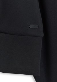 Tissu noir avec une couture subtile et une petite étiquette rectangulaire en métal noir embossée de "4F" près du bord.