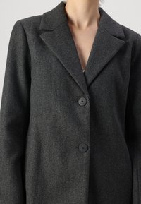 Šedý vlněný blazer s klopovým límcem, dvěma černými knoflíky a texturovaným povrchem. Přiléhavý střih, bez viditelných kapes.