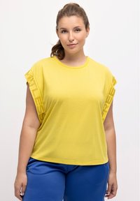 T-shirt giallo a maniche corte con scollatura arrotondata e stampa grafica lungo le maniche. Abbinato a pantaloni blu in tessuto testurizzato.