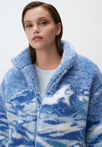 Giacca in pile blu caratterizzata da un motivo a onde testurizzato, con colletto alto e chiusura con zip. Materiale morbido e peloso e sfumature di blu variegate.
