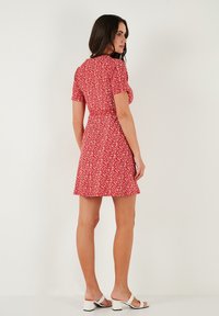 Rotes Kleid mit Blumendruck, kurzen Ärmeln, tailliertem Schnitt und schwingendem Rock. Glatter Stoff mit leichtem Tragegefühl, kombiniert mit weißen Sandalen.