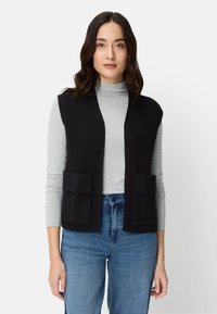 Frau mit dunklem, welligem Haar trägt eine schwarze, ärmellose Strickweste mit Taschen über einem hellgrauen Rollkragenpullover und blauer Jeans, steht neutral.