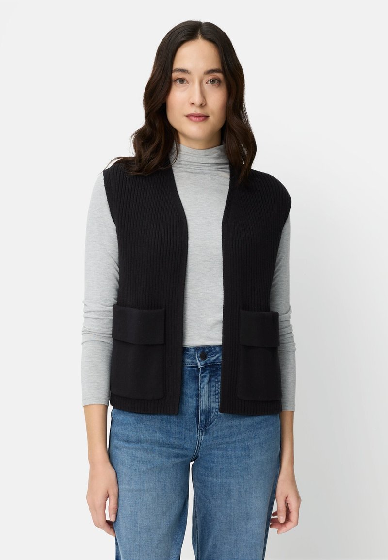 Frau mit dunklem, welligem Haar trägt eine schwarze, ärmellose Strickweste mit Taschen über einem hellgrauen Rollkragenpullover und blauer Jeans, steht neutral.