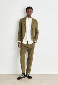 Olive green suit with a structured blazer and relaxed-fit trousers, paired with a white button-up shirt and black sneakers with white accents.