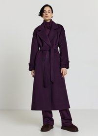 Cappotto lungo viola in lana con cintura in vita, rever ampi e polsini abbottonati, indossato sopra pantaloni viola abbinati e calzature scure.