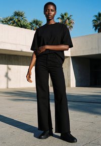 Kvinde med kort hår iført sort oversize t-shirt, sorte bukser med vide ben og sorte sneakers, stående udendørs med palmer og betonjbygninger.