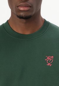 Dark green sweatshirt with a crew neck and ribbed cuffs, featuring a small pink embroidered bird design on the left chest.