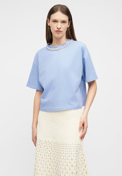 Γυναίκα που φοράει ανοιχτό μπλε κοντομάνικη μπλούζα με λεπτομέρεια από πέρλες στο ντεκολτέ και λευκή πλεκτή φούστα με διαμαντοειδείς ανοίγματα.
