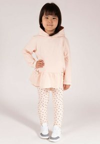 Jasnoróżowa sukienka dresowa z falbanką na dole, zestawiona z kremowymi legginsami w kropki. Buty białe z szarymi dodatkami. Całość wykonana z miękkiego, bawełnianego materiału.