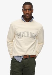 Sweat-shirt crème avec le logo "SUPERDRY" en gris sur la poitrine. Porté sur une chemise à col bleu clair. Pantalon vert olive, ceinture marron.