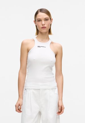 Młoda kobieta o prostych krótkich włosach nosi biały bezrękawnik z logo „KARL LAGERFELD JEANS” oraz białe luźne spodnie.