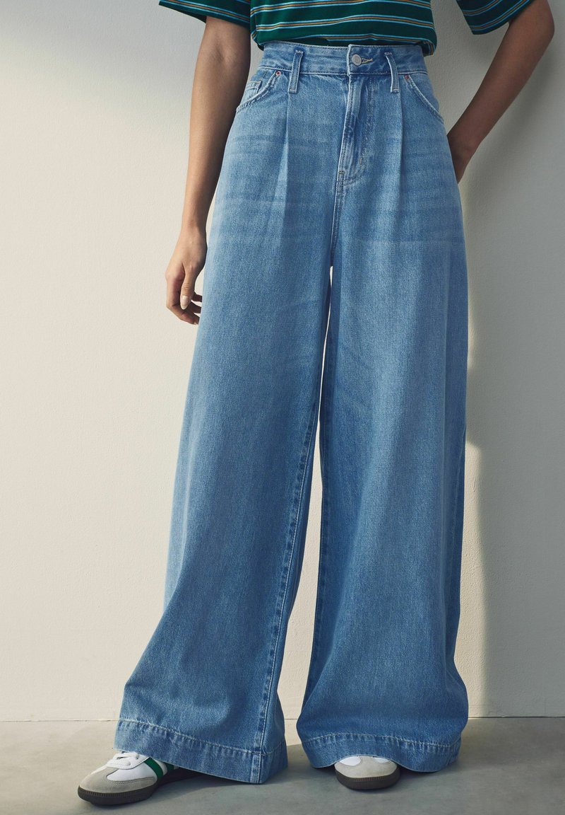 Pantalones de mezclilla amplios azul con cintura alta, que presentan pliegues frontales y un ajuste relajado, combinados con zapatillas blancas y grises.