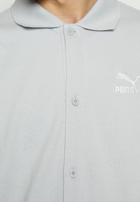 Polo gris clair en tissu texturé, doté d'un col, de deux boutons et d'un logo Puma blanc brodé sur le côté gauche.