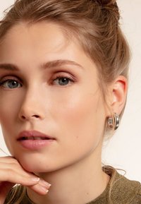 Silberne Creolen mit polierter Oberfläche, die ein klobiges Design und eine glatte Textur präsentieren. Die Hand des Modells ruht unter ihrem Kinn.