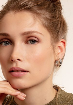 Silberne Creolen mit polierter Oberfläche, die ein klobiges Design und eine glatte Textur präsentieren. Die Hand des Modells ruht unter ihrem Kinn.