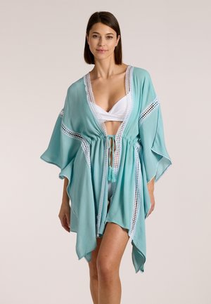 Frau trägt weißen Bikini und hellblauen Überwurf mit Spitzensaum, steht vor einfachem Hintergrund.