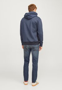 Sudadera azul marino con capucha, tejido mezclado, puños acanalados y un corte relajado, combinada con jeans azul oscuro y zapatillas de color claro.