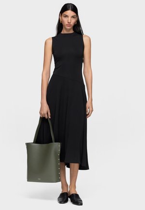 Woman with long dark hair wearing a sleeveless black dress, black slip-on shoes, gold hoop earrings, holding a large green tote bag with studs.