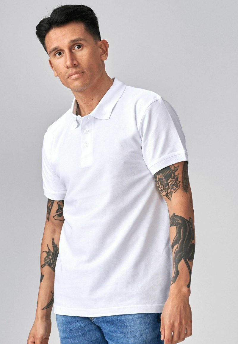 Homme aux cheveux noirs courts portant un polo blanc et un jean bleu, montrant des tatouages sur les deux avant-bras et regardant légèrement sur le côté.