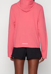 Sweat à capuche léger à manches longues en corail vif avec un col montant, doté de détails réfléchissants et d'une coupe décontractée. Associé à un short noir.