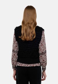 Gilet noir en tricot avec des volants texturés dans le dos, superposé sur une blouse à imprimé floral avec des manches longues et des poignets élastiques.
