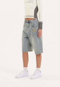 Pantalones cortos de mezclilla azul con un corte relajado, que presentan bolsillos frontales y un logotipo amarillo bordado. Combinados con zapatillas blancas y calcetines tobilleros.