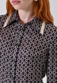 Giovane donna con capelli rossi che indossa una blusa nera con un motivo a catena chiaro e un dettaglio colletto beige, orecchino a cerchio argento visibile.