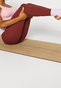 Bordeaux leggings gemaakt van rekbaar materiaal, met een aansluitend ontwerp en een gladde textuur. Een lichtroze sporttop is gedragen, tegen een houtkleurige achtergrond.