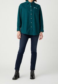 Camicia di velluto a coste verde acqua con bottoni, maniche lunghe, tasca sul petto e bottoni argentati, abbinata a jeans in denim scuro e stivali neri alla caviglia.