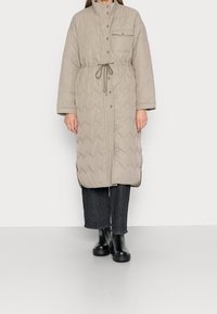 Abrigo largo de color beige acolchado con un alto cuello, botones a presión, bolsillo frontal, cintura ajustable con cordón y un patrón de acolchado en forma de chevrón. Se lleva sobre unos pantalones vaqueros de pierna ancha negros y botas.