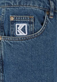 Plave traperice s leđa imaju džep sa izvezenim bijelim logotipom. Tkanina ima glatku teksturu i standardne šavove.