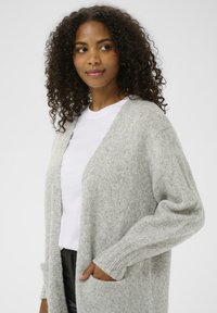 Kobieta z kręconymi włosami w jasnoszarym swetrze z dzianiny, nosząca białą koszulę, z rękami w kieszeniach, lekko patrzy w lewo.