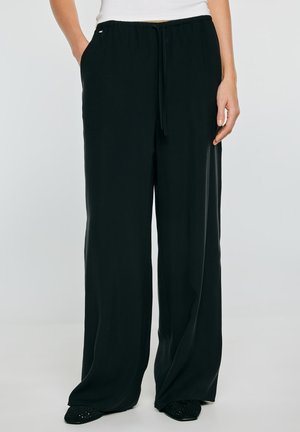 Kobieta nosząca luźne czarne spodnie ze ściągaczem w talii, ręce w kieszeniach, zestawione z czarnymi butami i białym skróconym topem do talii.