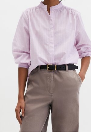 Personne portant un chemisier à manches longues rayé violet clair rentré dans un pantalon taille haute taupe avec une ceinture noire et une boucle dorée.