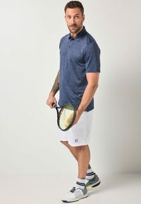 Sportlicher Mann in marineblauem Polo und weißen Shorts, der einen Tennisschläger und einen Ball hält, vor einem schlichten hellen Hintergrund stehend.