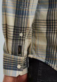 Chemise à manches longues, à carreaux en beige, bleu et noir. Présente des poignets boutonnés, des motifs contrastants et une petite étiquette de logo près de l'ourlet.