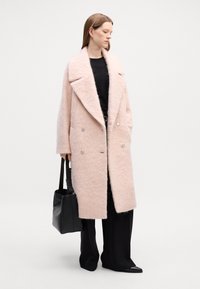 Casaco de mistura de lã rosa claro com um design de duplo botão, lapelas largas e bolsos laterais, combinado com uma bolsa preta e calças.