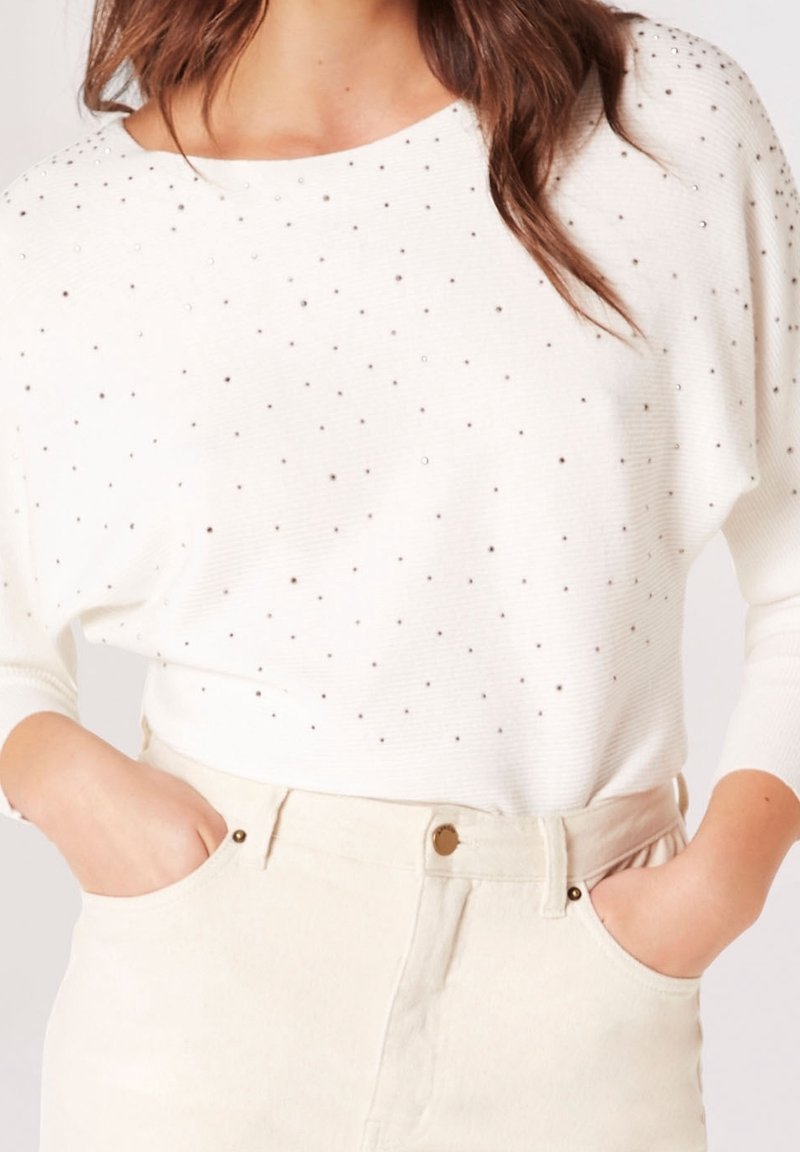 Femme portant un pull blanc avec de petits clous argentés et un pantalon taille haute écru, mains dans les poches.