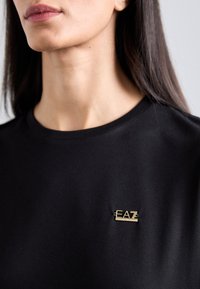 Juodas trumpomis rankovėmis marškinėliai su ryškiu auksiniu "EAZ" logotipo segtuku krūtinėje, pagaminti iš glotnaus audinio su paprastu apvaliu iškirpimu.