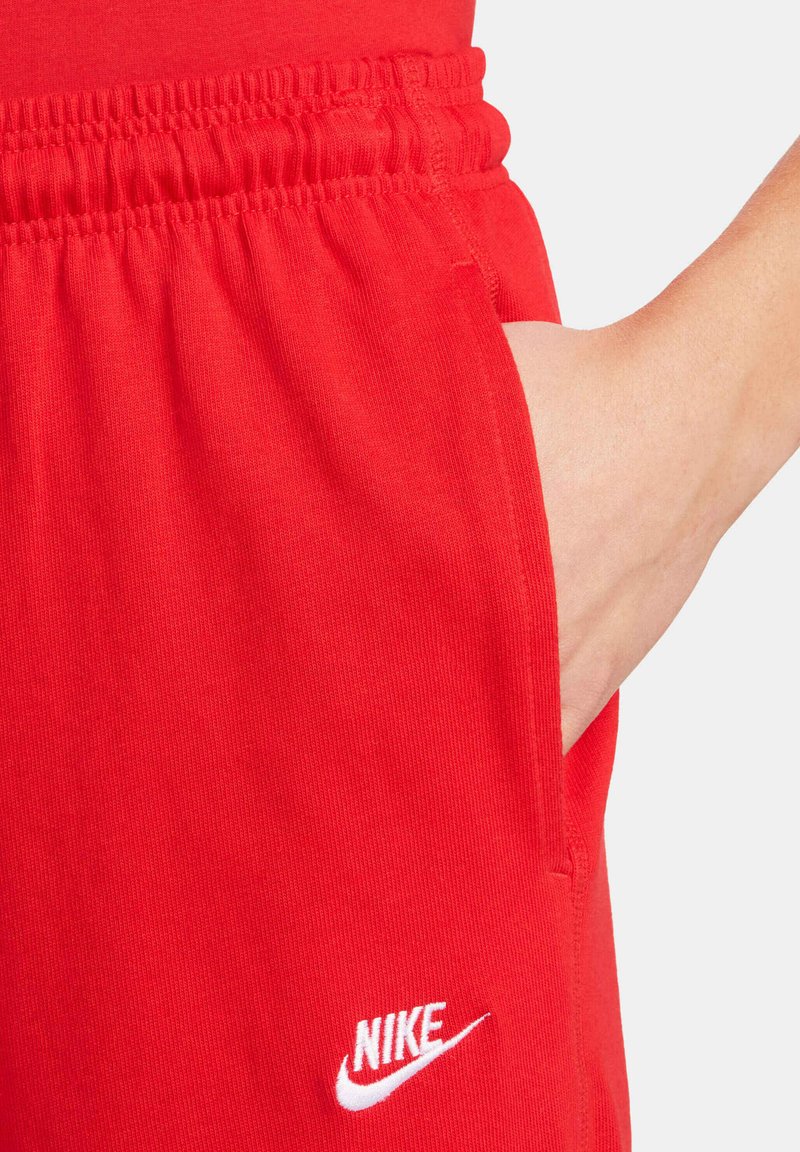 Czerwone dresy bawełniane z elastycznym paskiem w talii i białym logo Nike. Posiadają kieszenie boczne i gładką teksturę. Zbliżenie na materiał.