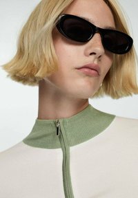 Tajta topp med halvdragkedja och grön ribbad krage samt krämfärgad kropp. Modellen bär svarta rektangulära solglasögon.