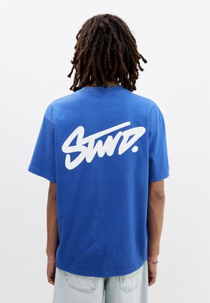 Blue cotton t-shirt featuring a large white graphic logo on the back. The shirt has short sleeves and a classic fit.