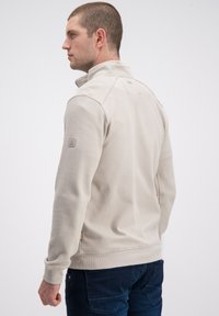 Jasnobeżowa bluza na zamek błyskawiczny z teksturowanej tkaniny, z wysokim kołnierzem i dopasowanym krojem. Posiada logo na rękawie oraz boczne kieszenie.