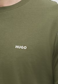 Βαμβακερό T-shirt ελιάς με στρογγυλή λαιμόκοψη και λευκό κείμενο "HUGO" τυπωμένο στο στήθος.