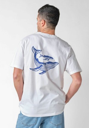Uomo che indossa una maglietta bianca con grafica di una balena blu e un piccolo sul retro, abbinata a jeans azzurri, in piedi davanti a uno sfondo semplice.