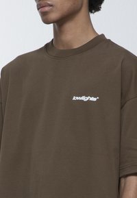 Camiseta de algodón marrón de gran tamaño con un logo blanco de "lowlights" en el pecho. Cuello redondo y mangas cortas, textura suave.